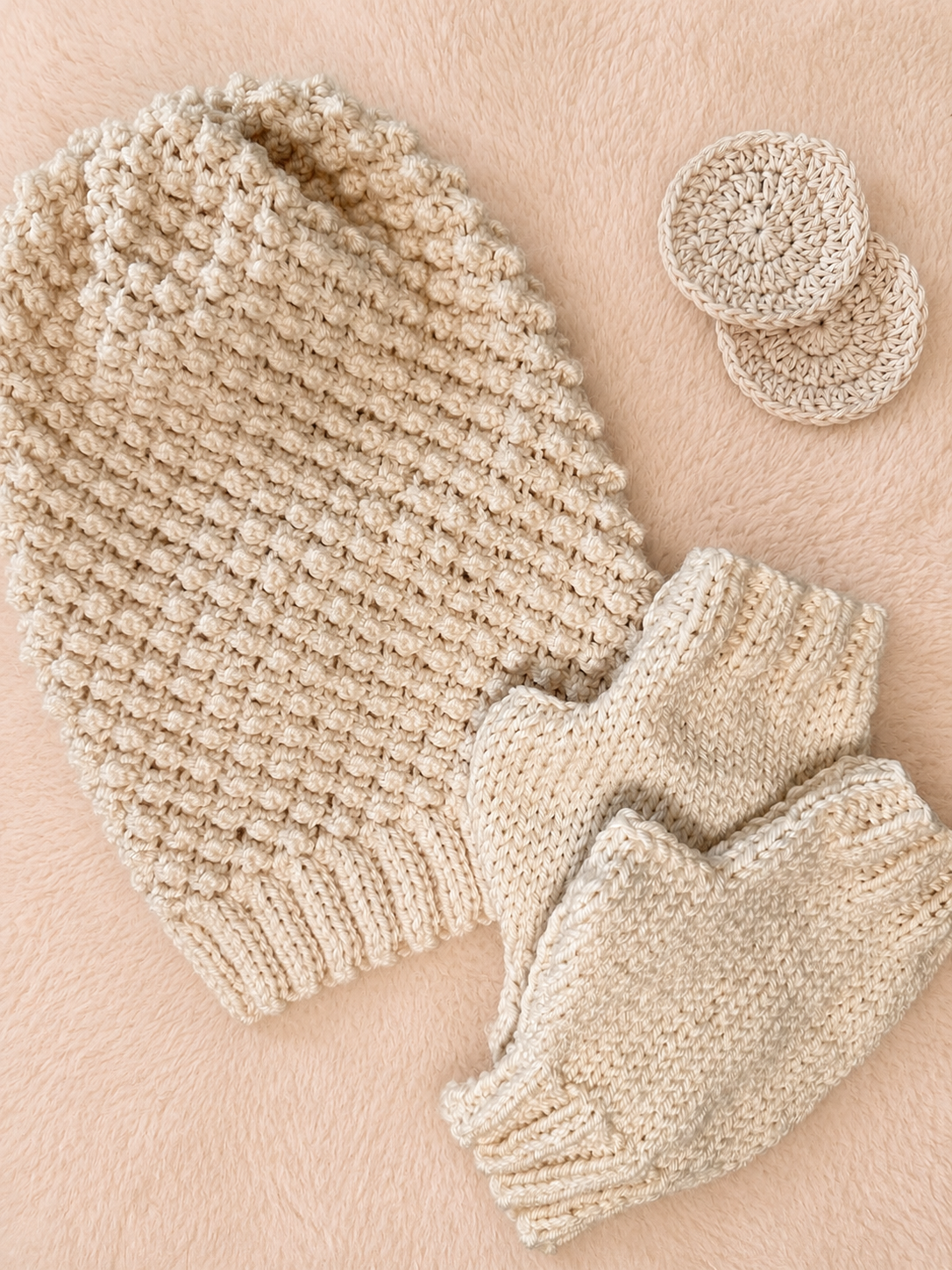 A beige blackberry stitch beanie with matching fingerless gloves and two beige face scrubbies, arranged on a fluffy background.,