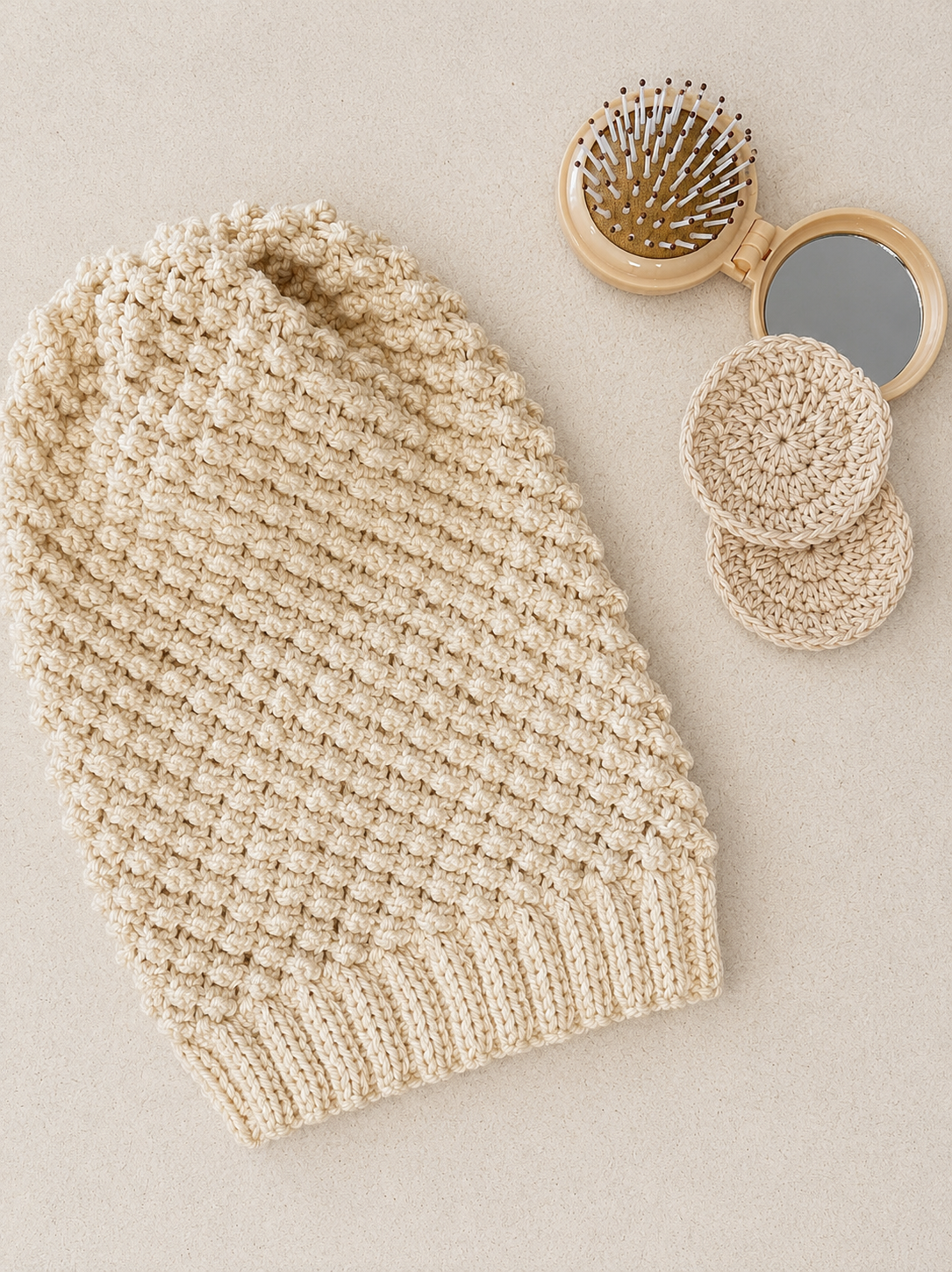 A flat lay of a beige blackberry stitch slouch beanie next to two crocheted face scrubbies and a mirror and hairbrush.