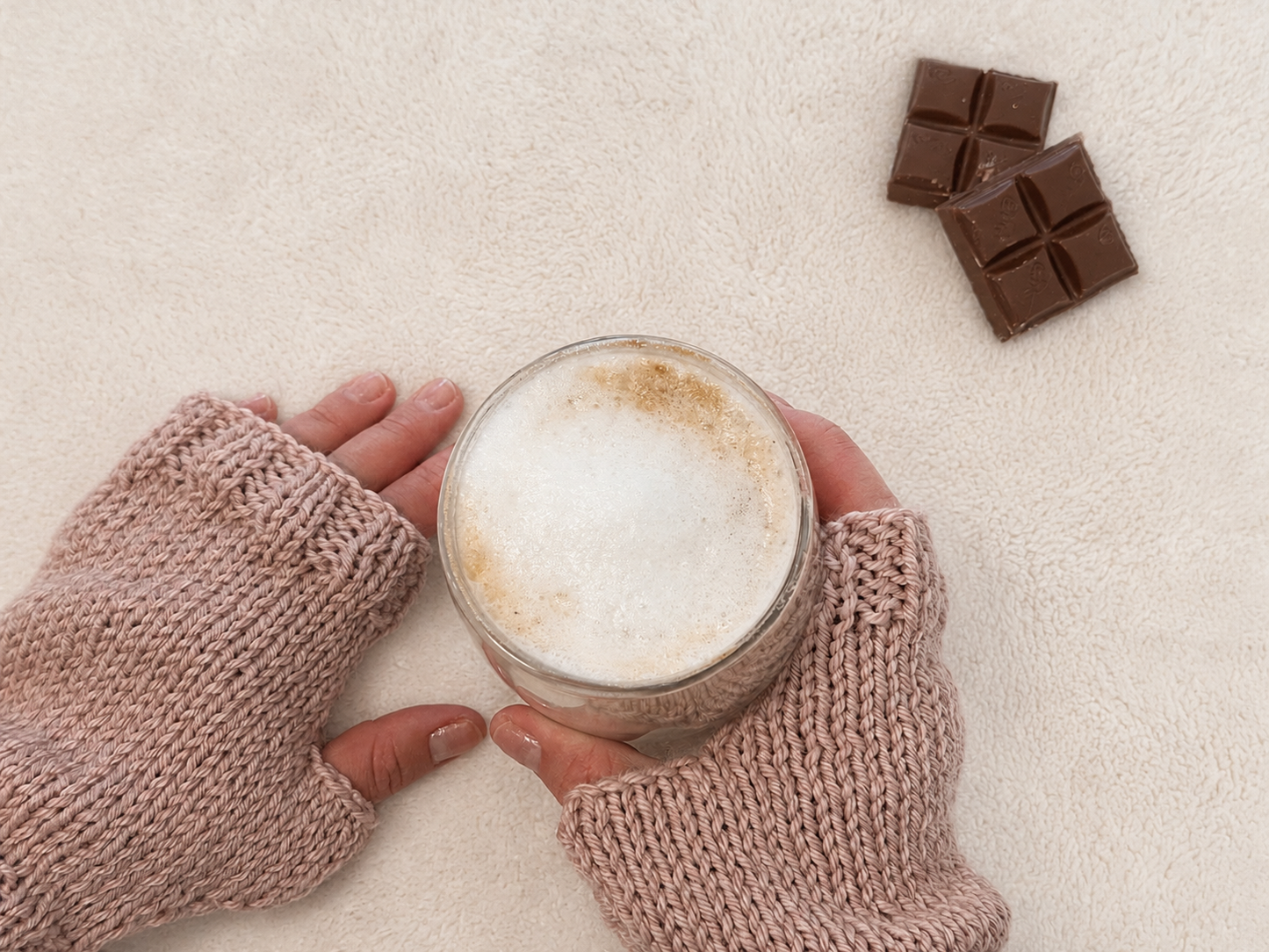 A top-down view of a person's hands wearing pink knit fingerless mittens holding a glass of hot chocolate with thick foam, with two chocolate bars on a textured cream background.