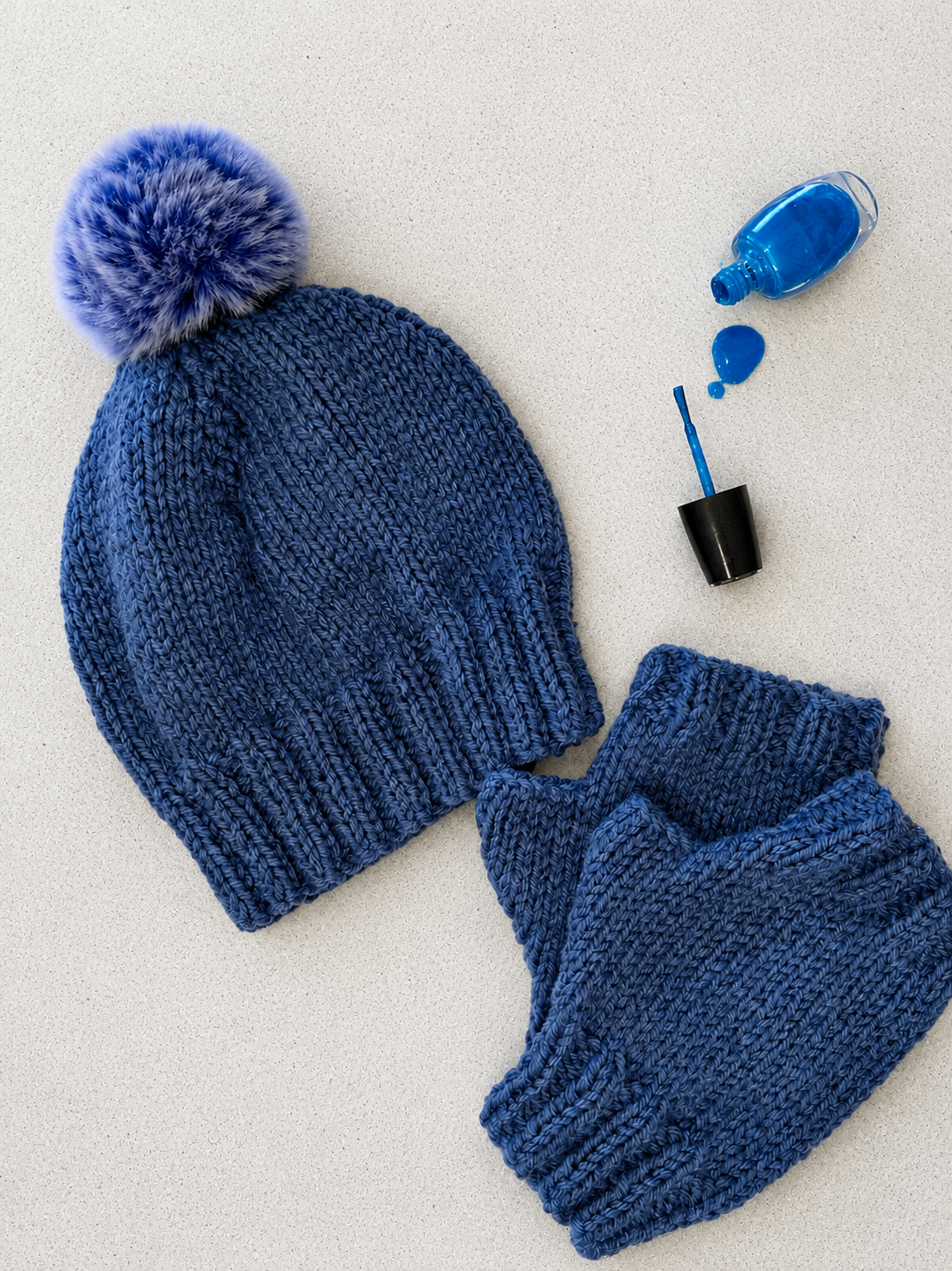 A blue knit beanie with a fluffy pom-pom and matching knit mittens, styled on a light textured background with a small bottle of bright blue polish and an applicator brush nearby.