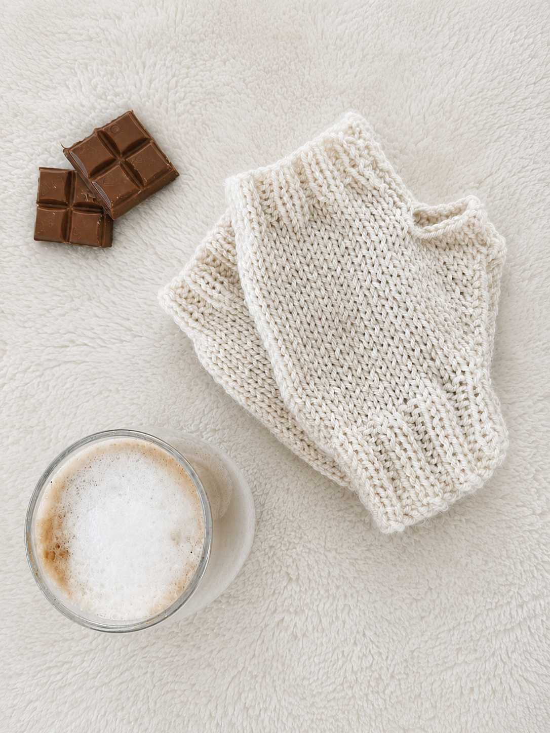 A pair of beige, knit fingerless gloves laid flat on a soft white textured surface, styled with two chocolate bars and a glass of frothy coffee