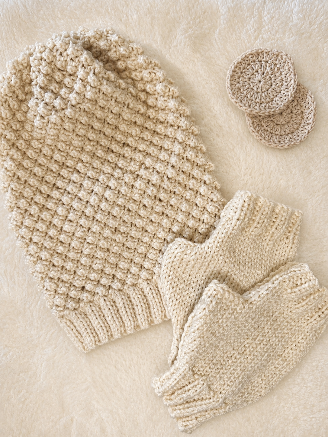 A beige coloured knitted slouch hat flat lay, with a textured beanie, fingerless gloves, and two small round hats, arranged on a soft cream faux fur background 