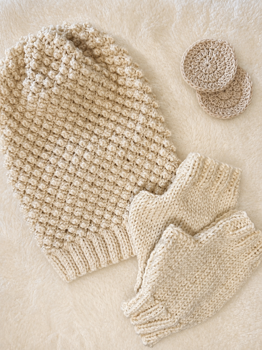 A beige coloured knitted slouch hat flat lay, with a textured beanie, fingerless gloves, and two small round hats, arranged on a soft cream faux fur background 