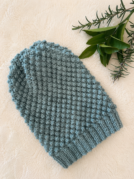 A dusty green textured blackberry stitch beanie laid flat on a cream-colored surface, styled with green leaves in the top right corner.