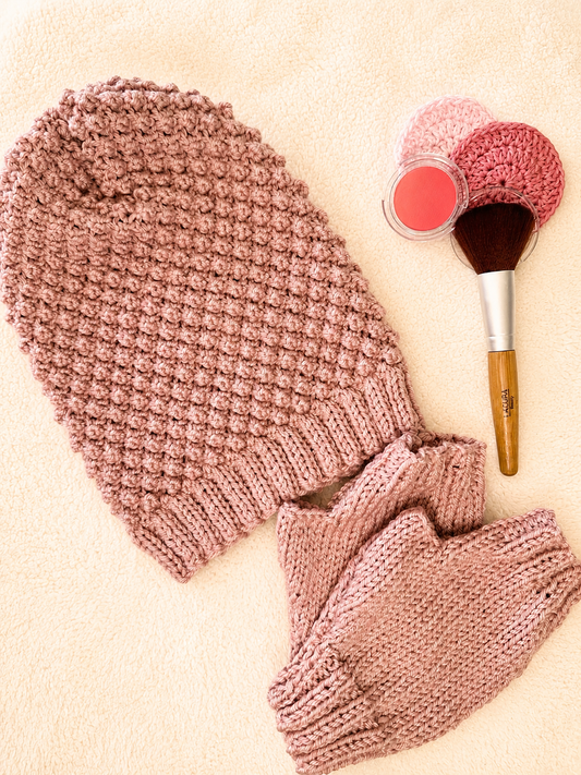 A dusty pink blackberry stitch slouch beanie and matching fingerless gloves laid flat on a cream fabric background, styled with a makeup brush, a blush compact and two pink face scrubbies.