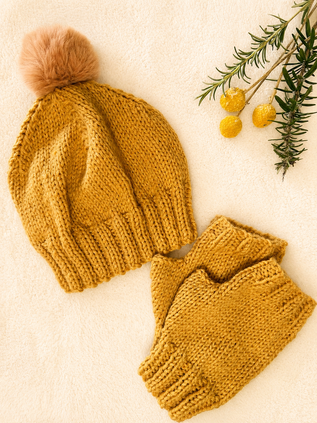 A flat lay of a cosy knit beanie and matching fingerless gloves in a warm mustard yellow, styled on a textured cream background with a brown pom-pom alongside small decorative yellow billy button flowers and rosemary.