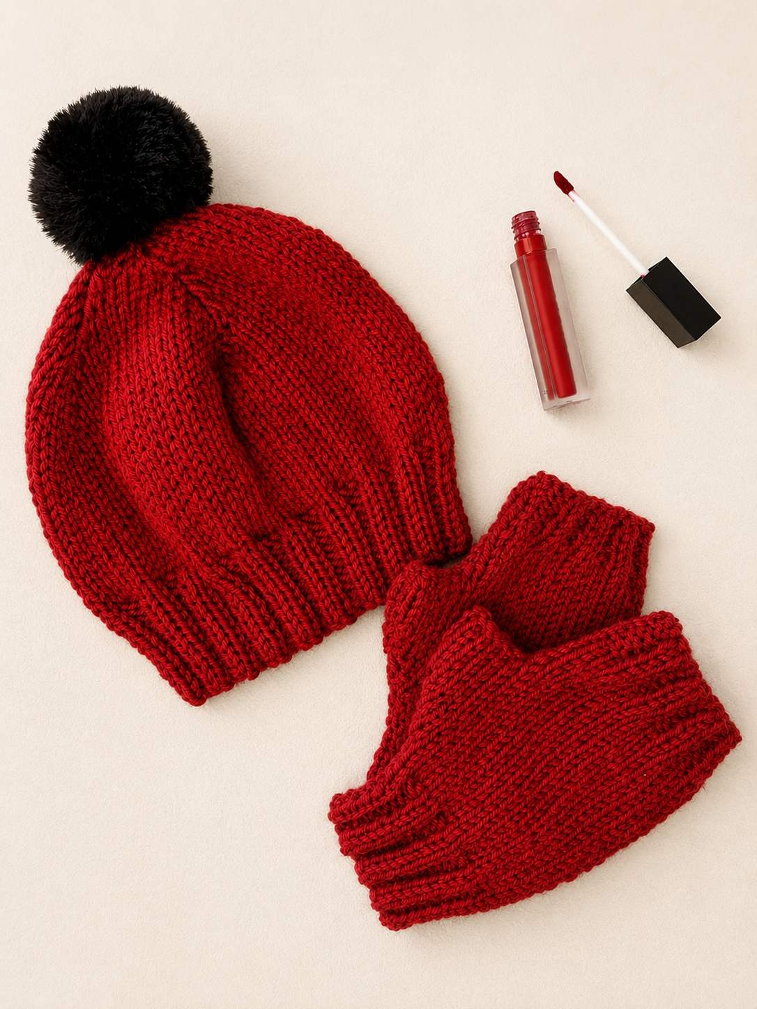 This image is of a flat-lay of a red beanie with a black pom pom, next to red fingerless gloves and a red lip gloss applicator