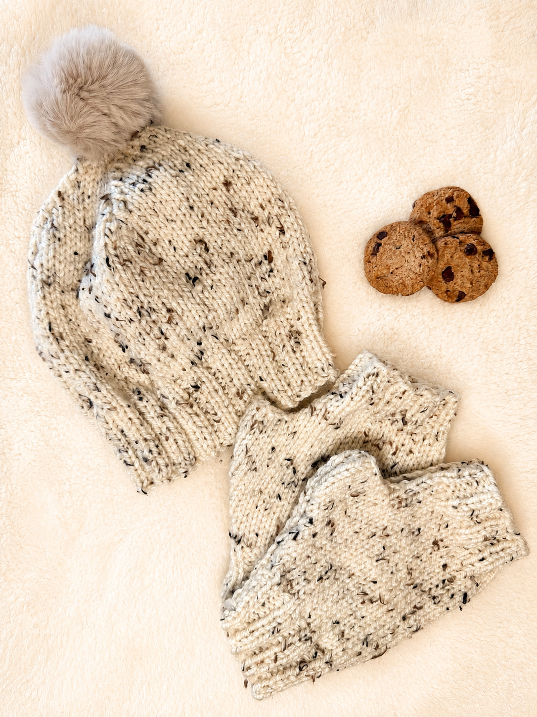 A knit beanie with a fluffy pom-pom and matching knit mittens in a cream-colored yarn with black and gray speckles, arranged on a soft cream background next to three chocolate chip cookies.